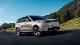 En sølvfarvet Jaeceon J5 EV SUV kører på en snoet vej gennem et naturskønt, bjergrigt landskab under en klar blå himmel.